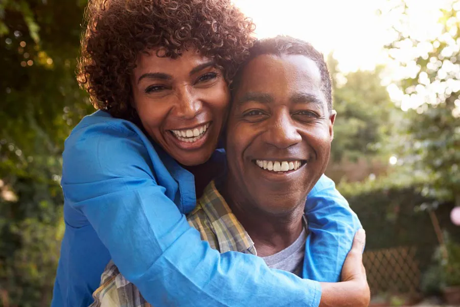 Autoimmune-Doctor A middle-aged African American couple outside, the woman embracing the man from behind pressing her cheek to his, both are smiling due to successful Autoimmune Disorder treatment from Stephen Beyer, DC in Mokena.
