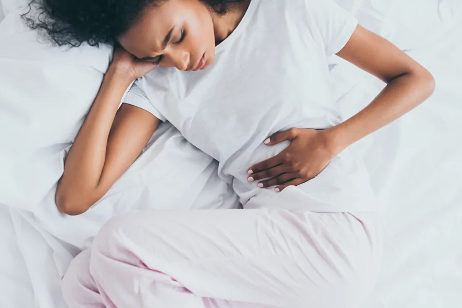 Crohns-Disease-Clinic A dark-skinned woman in white clothes lays on her bed clutching her middle before getting treatment for Crohn's Disease from Stephen Beyer, DC in Mokena.