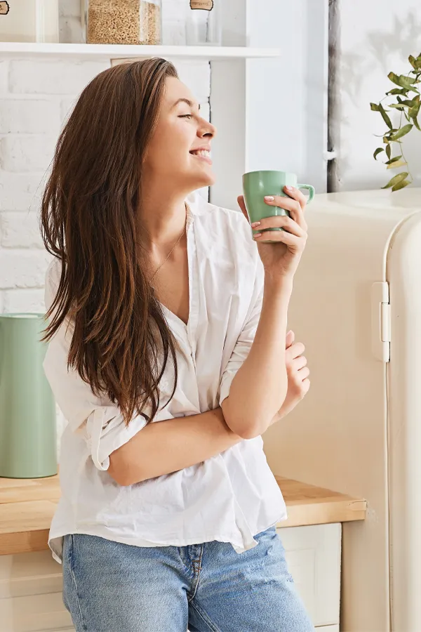 Crohns-Disease-Treatment A woman with a white blouse an jeans drinking tea in her kitchen, smiling after getting treatment for Crohn's Disease from Stephen Beyer, DC in Mokena.