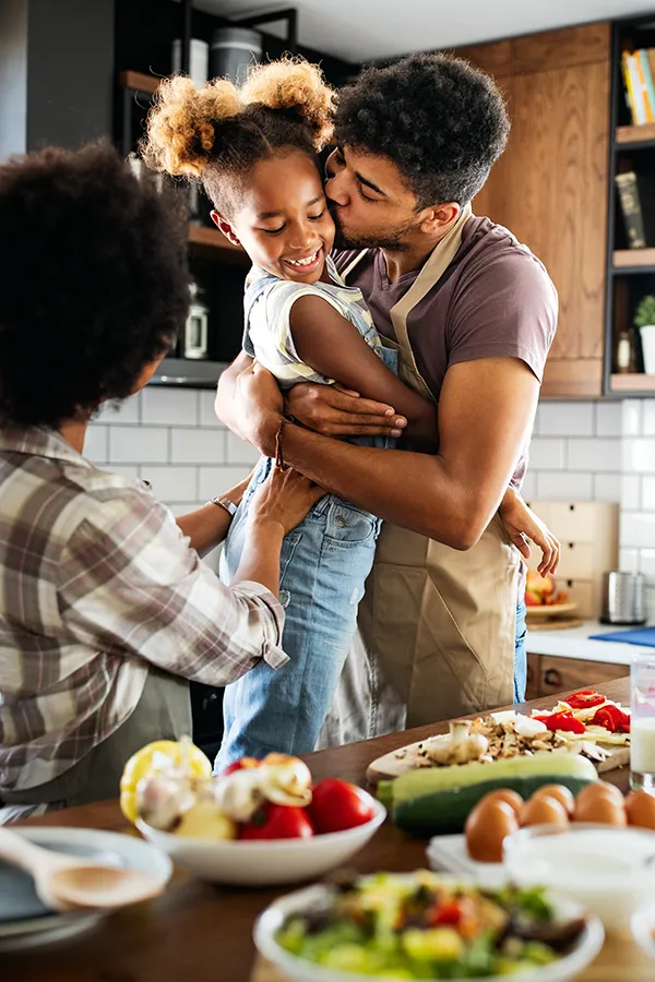 Gut-Health-Treatment A man holds his daughter and kisses her cheek as the family makes a healthy dinner in the kitchen. Get help for digestive issues and gut health from Stephen Beyer, DC in Mokena.