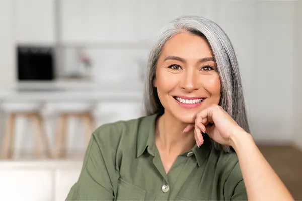 Menopause-Clinic An asian woman with long gray hair and a green shirt sitting and smiling on her couch, happy with her menopause care from [NAME} in Mokena.